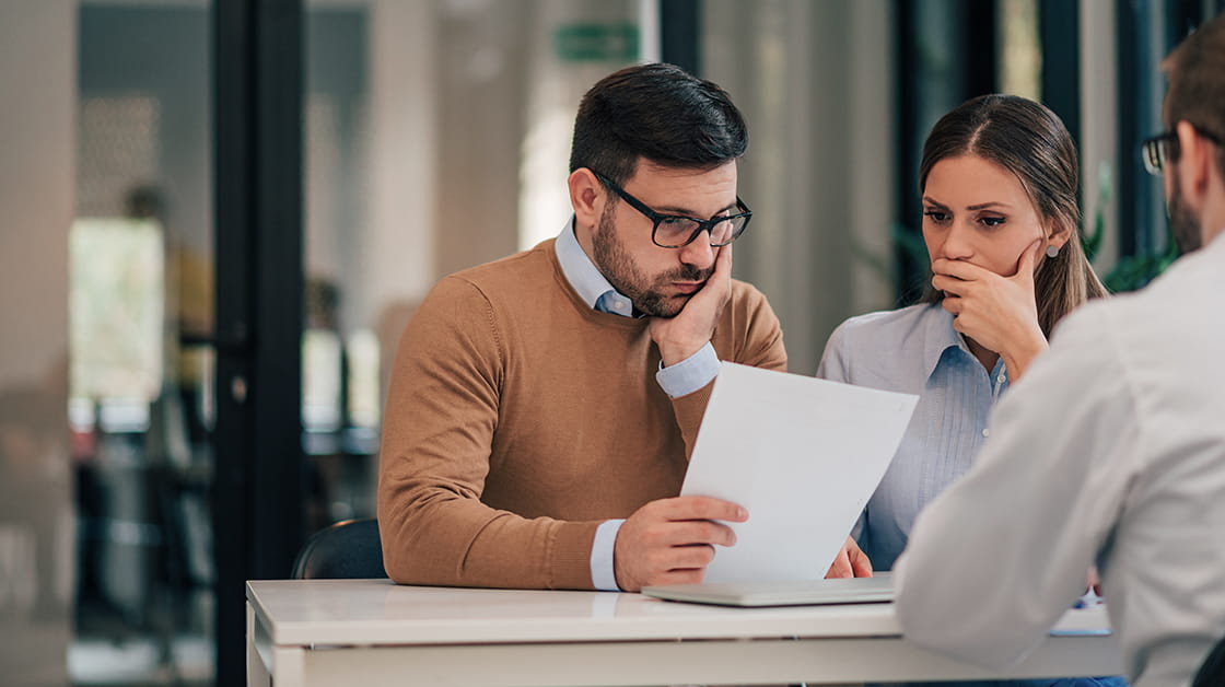 Two stressed clients sit across a desk from their solicitor. A white man with short dark hair and a beard is looking at paperwork with a concerned expression. His partner, a white woman with long, straight brown hair, looks similarly concerned.