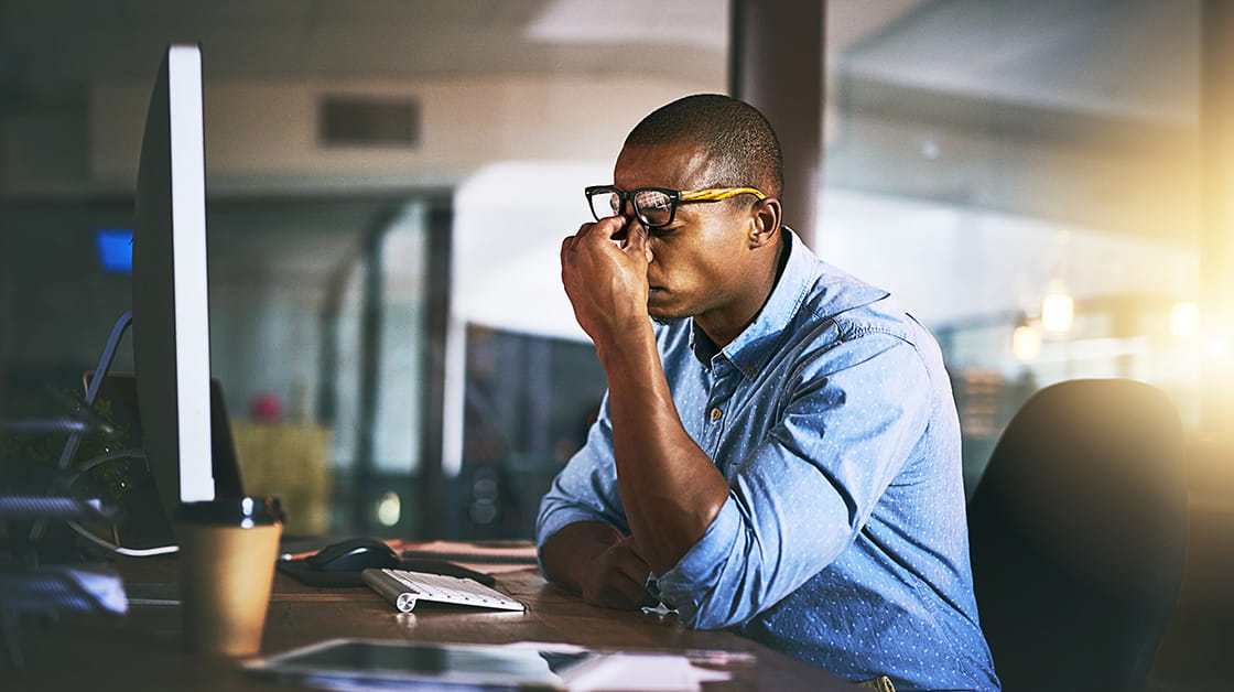 A stressed Black man with short black hair sits at a desk late at night with his head in his hands. He wears glasses and a blue shirt.