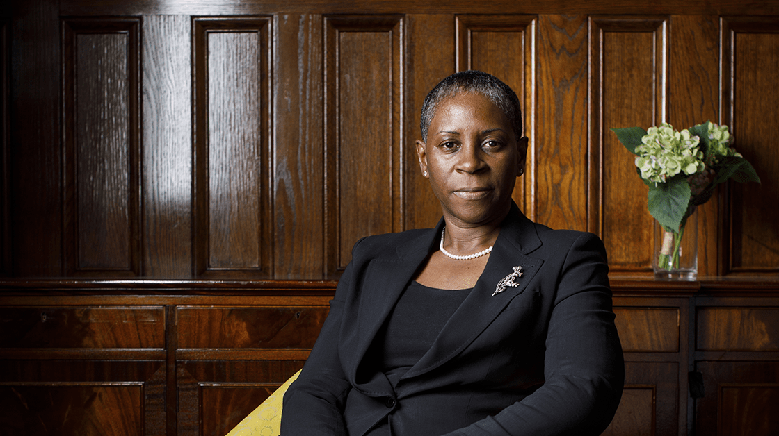I. Stephanie Boyce is a Black woman with cropped dark grey hair. She sits looking at the camera, wearing a black suit with a silver broach.