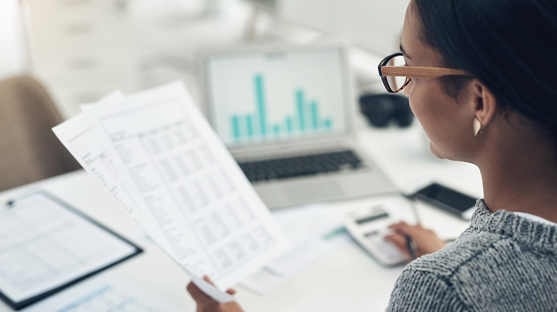 A black woman wearing glasses and a hair wrap sits at a desk, looking at finance documents and charts.