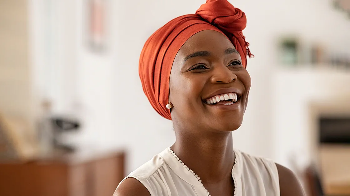 A black woman wearing an orange headscarf smiles widely. She stands in an office and wears a white blouse and pearl earrings.