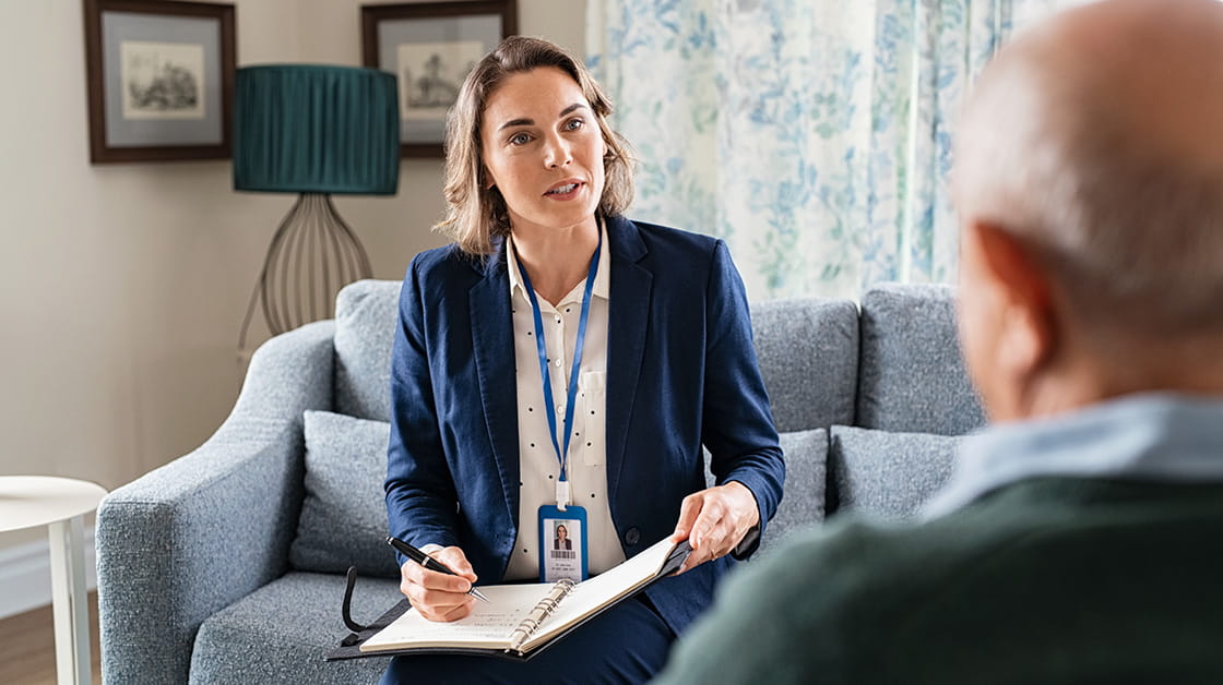 A professional woman with short, wavy dark blonde hair sits across from a balding elderly male client with grey hair. She wears a navy suit, white shirt and lanyard. He wears a green jumper and a blue shirt.