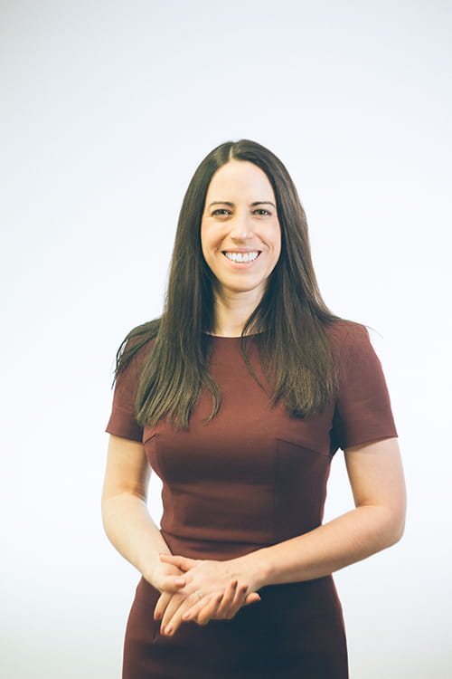 Deidre Sheahan is a white woman with long, dark-brown hair, wearing a red-brown dress, smiling at the camera