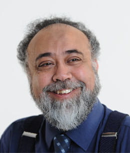 Faizal Essat is a man, wearing braces and a blue shirt and tie. He has a trimmed grey beard and smiles off as he looks to the left of the camera.
