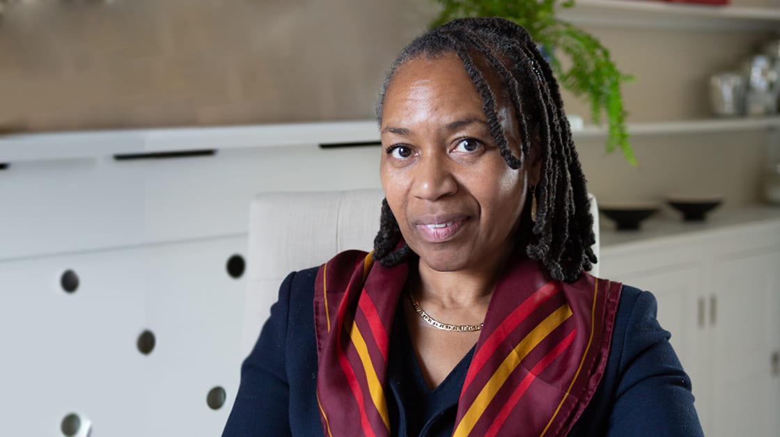 Jacqueline Devonish is a black woman with shoulder-length braids, wearing a red and gold silk scarf. She is looking at the camera, with a neutral living room in the background