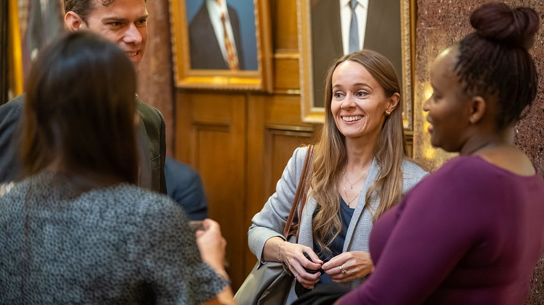 A white woman with long brown hair, wearing a grey jacket is smiling and chatting to three other people, including a Black woman with a purple top, a woman with brown hair and a black jacket and a man with short brown hair.