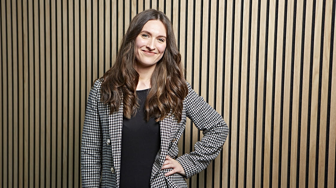Rebekah Sutcliffe is a white woman with long, wavy brown hair. She is smiling and wears a houndstooth suit over a black blouse.