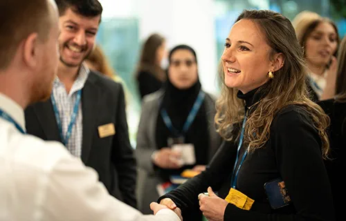 A white woman with brown curly hair is smiling and shaking hands with a man in a white shirt. In the background there are more people including a man with brown hair and a bear who is smiling at the two shaking hands. They are all wearing blue lanyards.