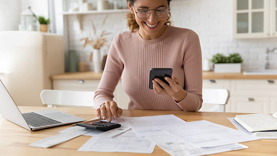 Smiling young woman wearing glasses,  using phone and calculator to work out insurance, budget or expenses, sitting at kitchen table with papers
