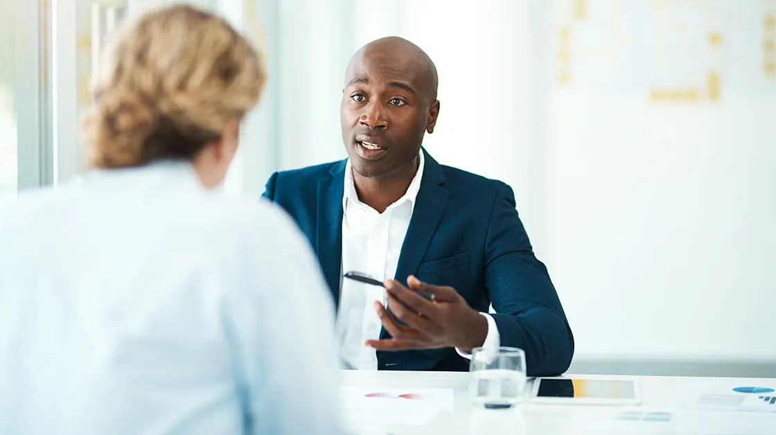 A Black man with closely cropped hair sits across a table from his white, female client with blonde hair. He wears a navy suit and white shirt. She wears a pale blue shirt.