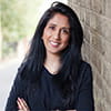 Anita is a woman with black hair and is wearing a black jumper. She is smiling into the camera and standing next to a brick wall.