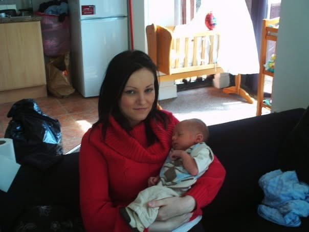 Claire Liddy in a red top holding a hew newborn son whilst sitting on a sofa with a crib in the background.
