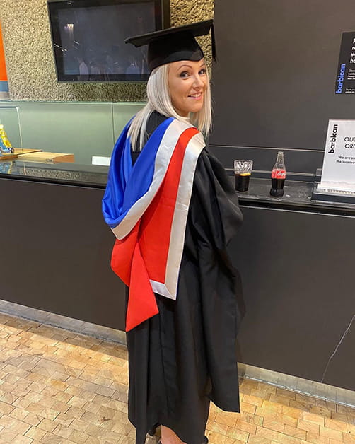 Claire Liddy smiles looking over her shoulder whilst wearing a graduation gown and cap at her masters graduation. Claire has blonde hair and is standing in a kitchen.