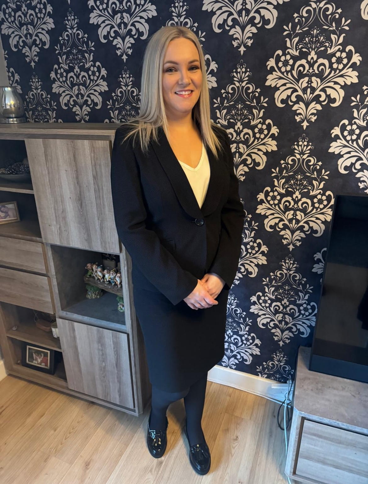 Solicitor Claire Liddy smiles wearing a smart black suit, she has blonde straight hair and stands in her living room in front of dark blue and silver patterned wallpaper.