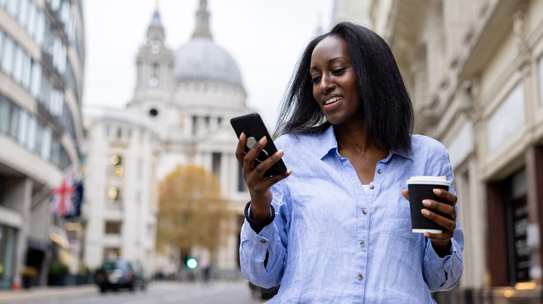 A young woman in business dress, walks down Ludgate Hill, London, holding a coffee and looking at her phone. 