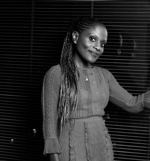 Solicitor MaameYaa Kwafo-Akoto smiles whilst standing in front of window blinds. The image is in black and white. MaameYaa is a black woman with long dark hair.