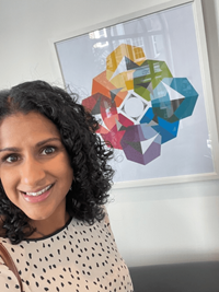 Neha is a woman with shoulder length, curly black hair. The image is a selfie, with a picture frame on the wall in the background.