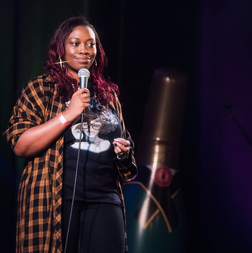 Sikisa Bostwick-Barnes stands on stage with a microphone performing a stand-up routine at a comedy club. She wears a baggy t-shirt and checked shirt over the top. She has black hair with red highlights.