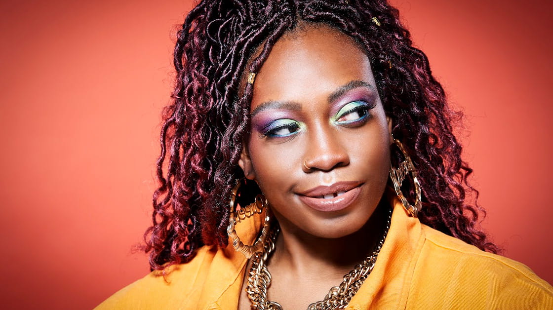 Comedian Sikisa stands in front of a dark orange backdrop whilst wearing an orange shirt. Her smile is a wry one and she looks off to the side with an air of intrigue. She has black curly hair with red highlights.