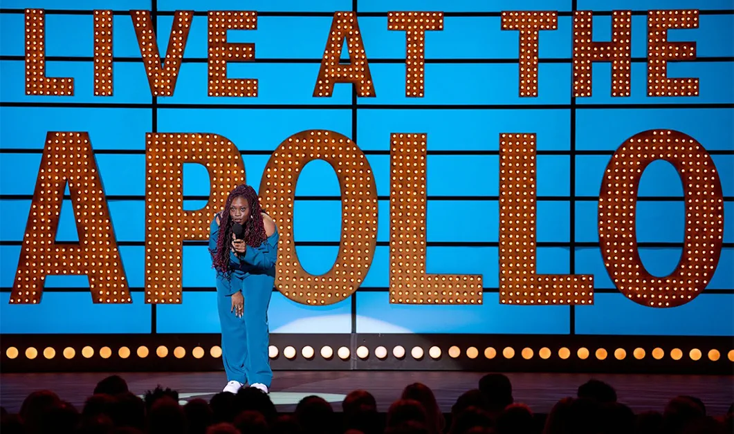 Sikisa Bostwick-Barnes stands on stage in front of an audience holding a microphone, wearing a bright blue outfit. She is in front of a Live At the Apollo backdrop and is performing comedy.