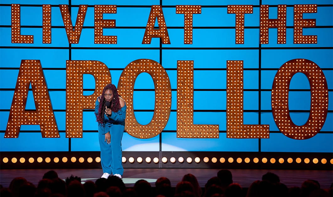 Sikisa Bostwick-Barnes stands on stage in front of an audience holding a microphone, wearing a bright blue outfit. She is in front of a Live At the Apollo backdrop and is performing comedy.