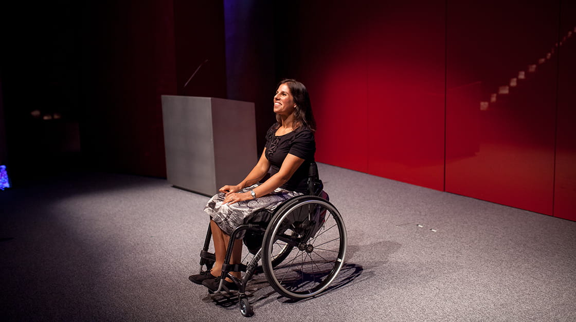 Yasmin Sheikh is a mixed-race woman with medium-length, wavy, dark brown hair. She is smiling widely and sits in a wheelchair on a stage, addressing an audience. She wears a black t-shirt, a silver and black patterned skirt and black shoes.