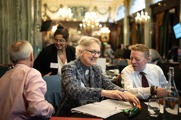 Four people seated around a table in a formal room with chandeliers and large windows, engaged in discussion with papers and a water bottle on the table.