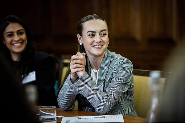 A woman with long hair tied back, wearing a grey plaid blazer, sits at a table with hands clasped. She is smiling, with papers, a pen, and a glass of water in front of her. 