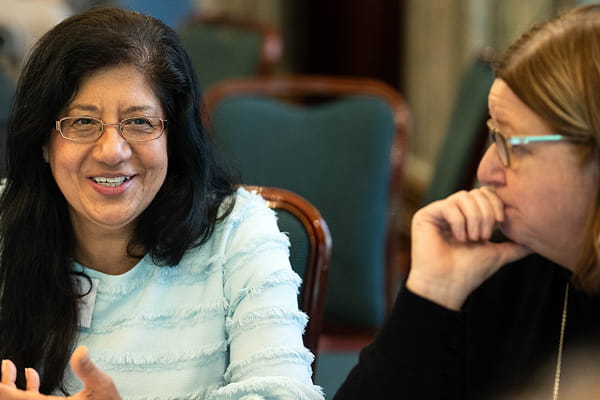Two people seated and talking in a room with green chairs; one smiles while speaking, the other listens thoughtfully with their chin resting on their hand.