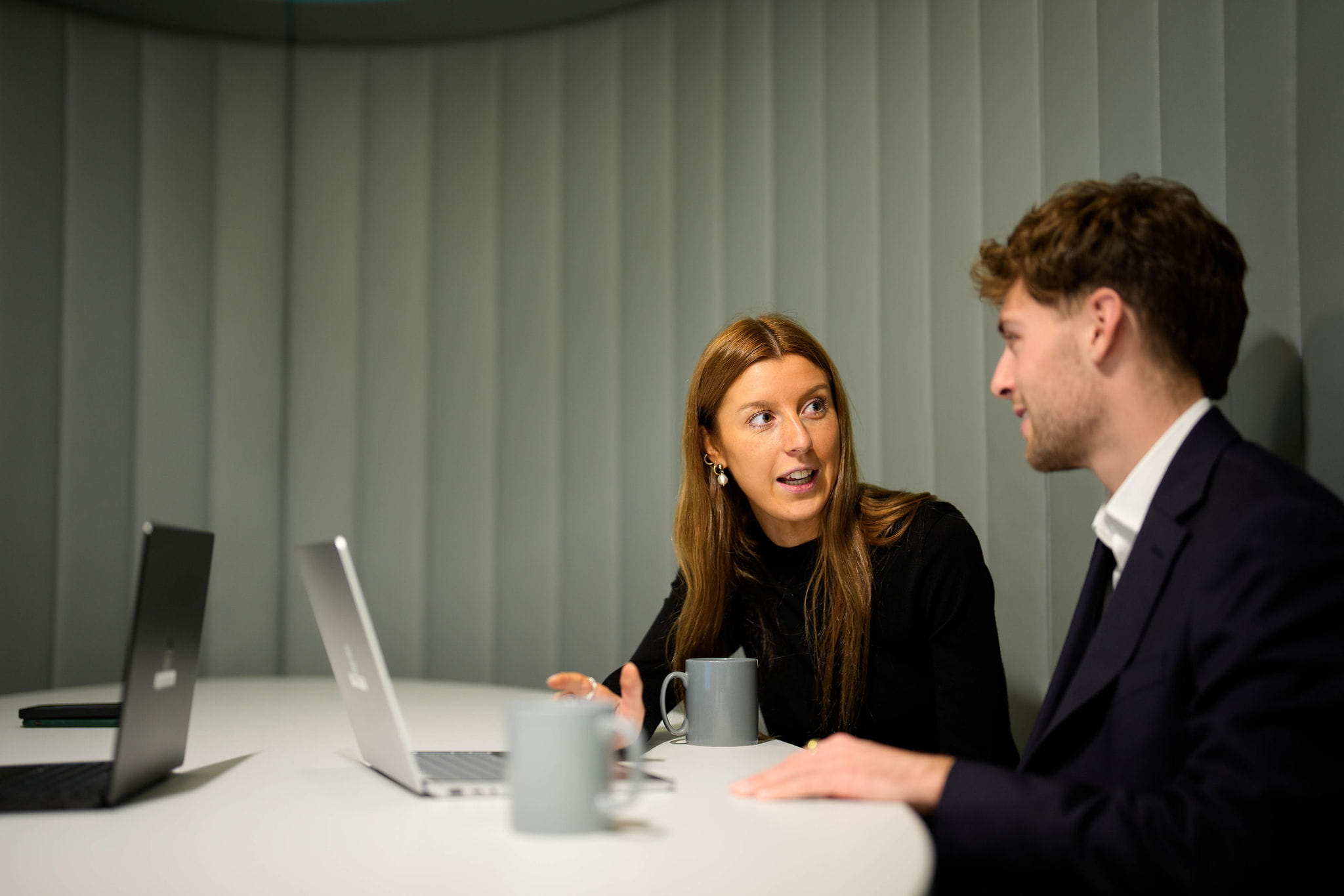 two people sitting at a table and talking. one is wearing a suit, the other is in a black jumper.